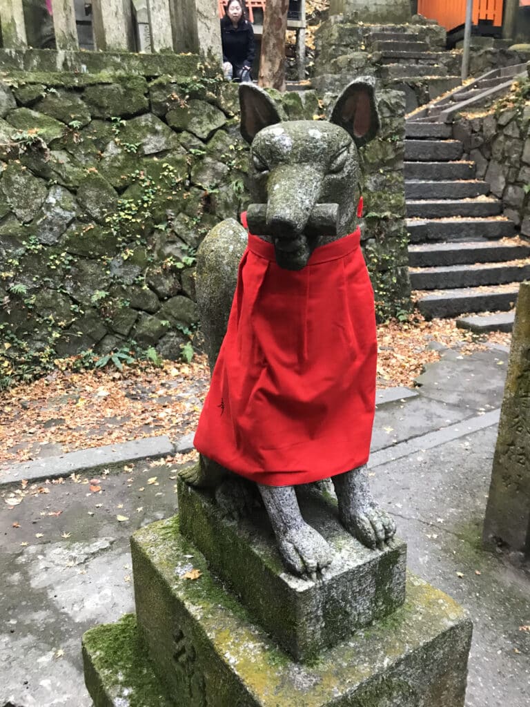 A kitsune statue wearing a red bib while holding a scroll in its mouth at the Fushimi Inari Shrine