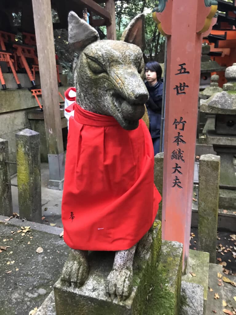 A kitsune statue wearing a red bib while holding a ball in its mouth at the Fushimi Inari Shrine