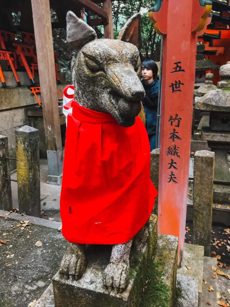 Kitsune fox messenger statue wearing a red votive bib at Fushimi Inari Taisha, Kyoto, with a ball in its mouth