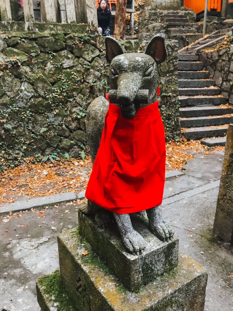 Kitsune fox messenger statue wearing a red votive bib at Fushimi Inari Taisha, Kyoto, with a scroll in its mouth