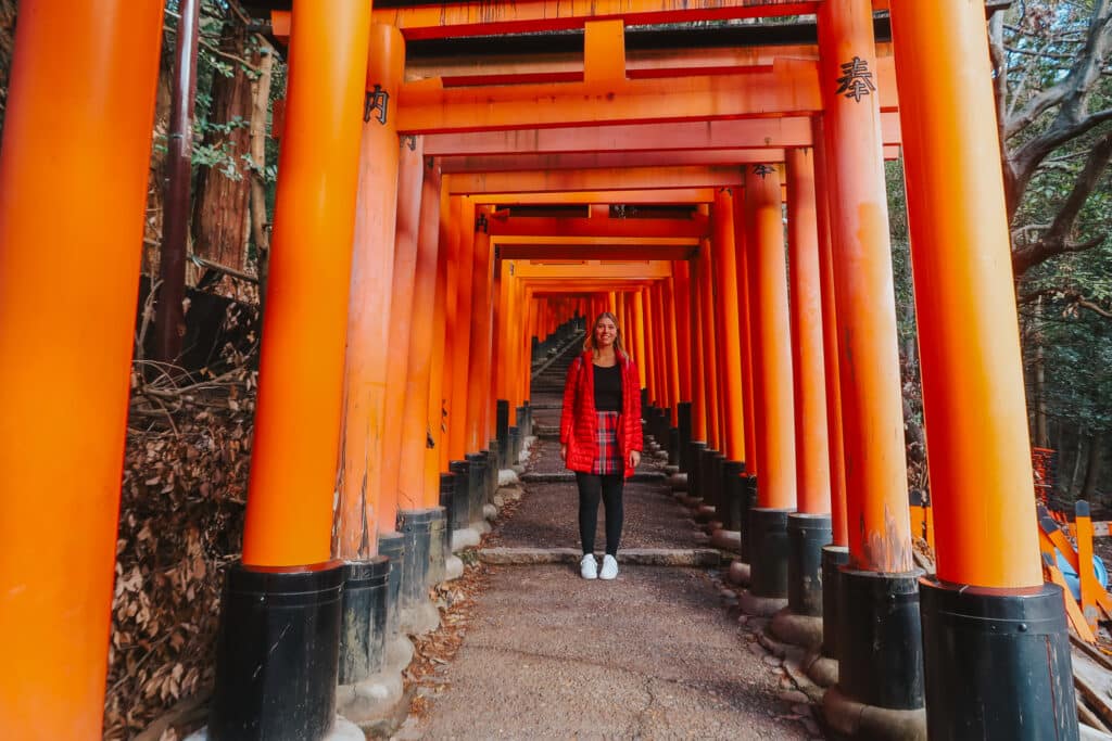 Hiker under a vermillion Torii gate tunnel lined with autumn leaves at Fushimi Inari Taisha, Kyoto