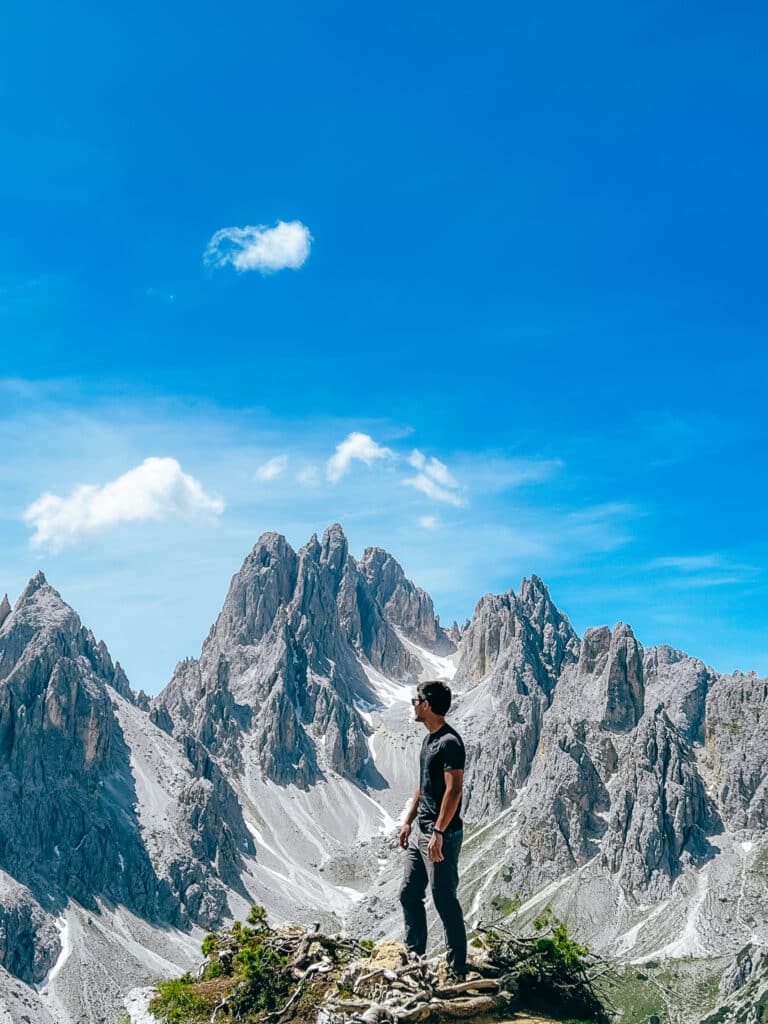 Male hiker in a grey shirt standing on a rocky ledge below the dramatic Cadini di Misurina spires in the Italian Dolomites