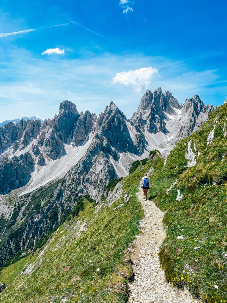 Solo hiker approaching the Cadini di Misurina viewpoint on a gravel path edged with wildflowers in the Dolomites, Italy