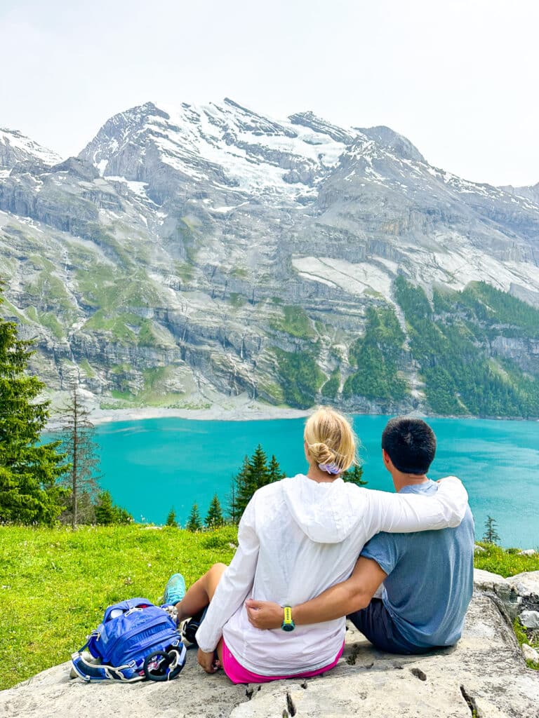 Couple sitting together on a rock overlooking Oeschinensee lake from behind, arms around each other, Swiss Alps ahead