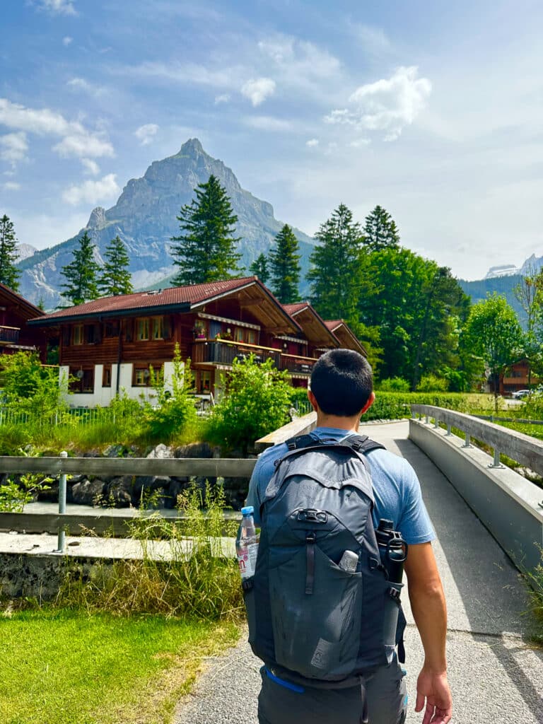 Hiker with backpack walking through Kandersteg village toward a dramatic rocky mountain peak in summer, Switzerland