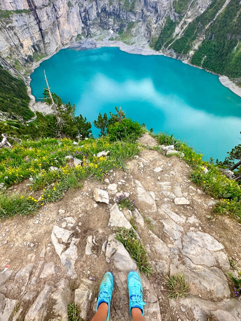 Hiker's feet in blue trail shoes on a rocky ledge with Oeschinensee's turquoise lake and wildflowers far below