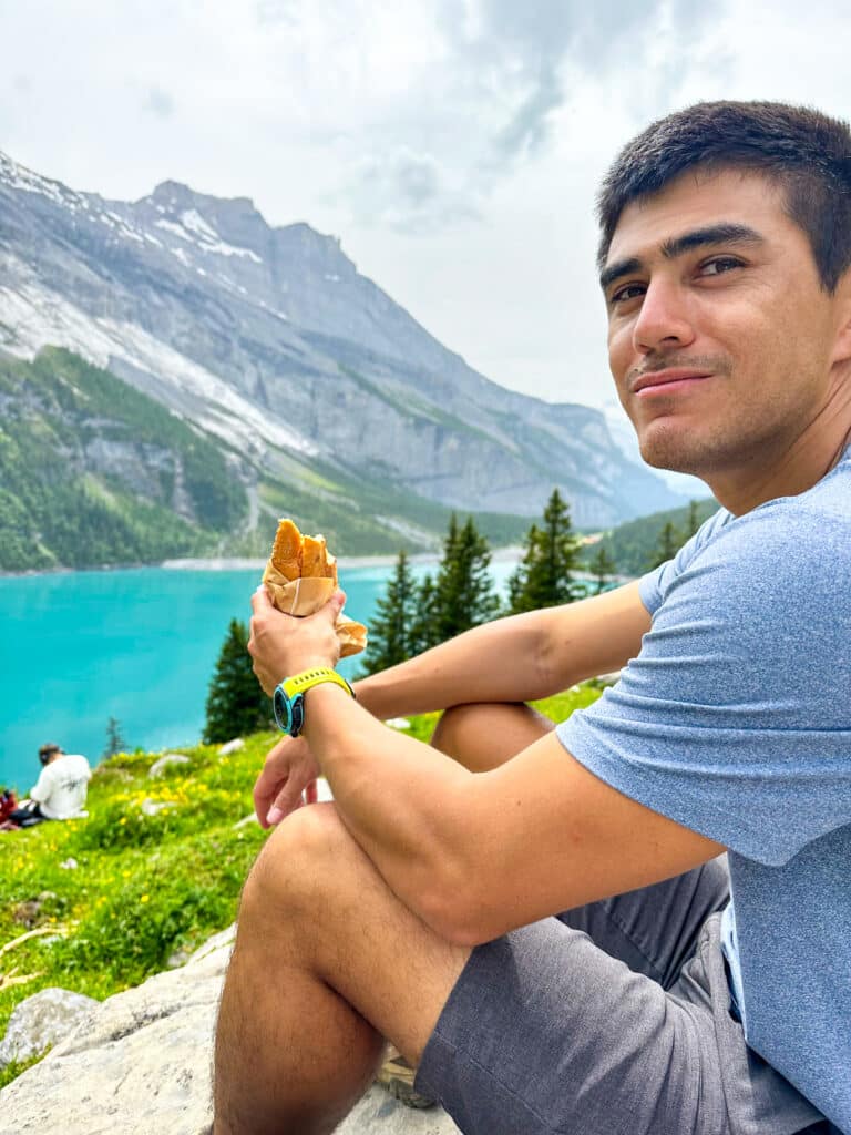 Man eating an sandwich at a rest stop beside Oeschinensee lake with turquoise water and mountains behind