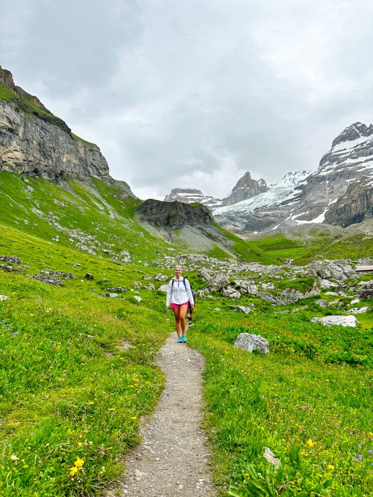 Woman hiking away on a green mountain trail above Oeschinensee with a sweeping valley and cloud-covered peaks ahead