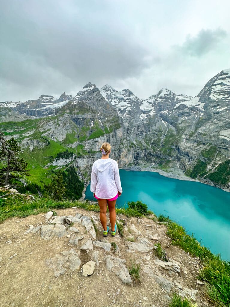 Woman overlooking Oeschinensee's turquoise lake from a rocky ridge, surrounded by snow-capped Swiss mountain peaks