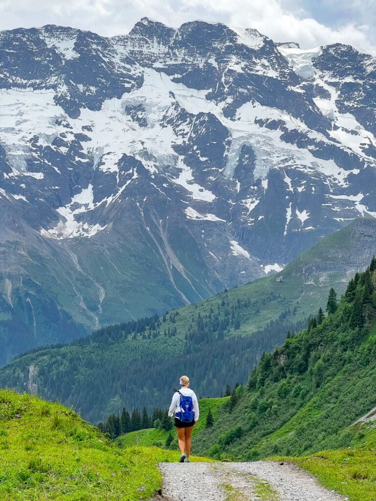 Female hiker with a blue daypack pausing to take in a vast wall of snow and rock on the North Face Trail above Mürren, Switzerland