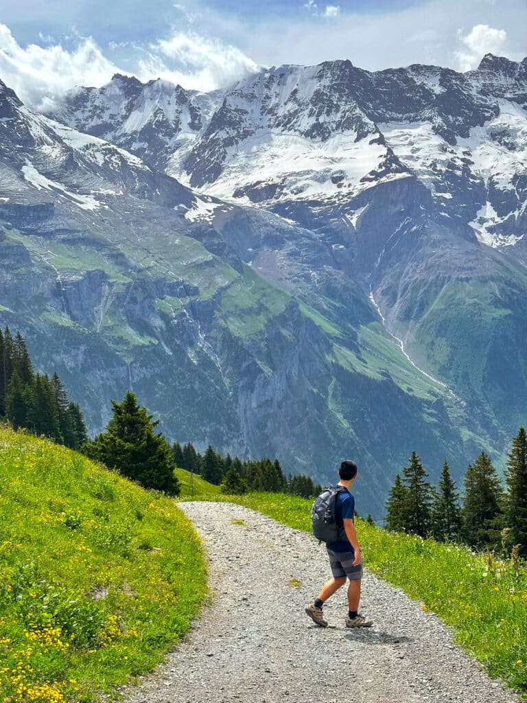 Male hiker with a daypack walking a gravel mountain path toward snow-covered peaks on the Northface Trail near Mürren, Switzerland