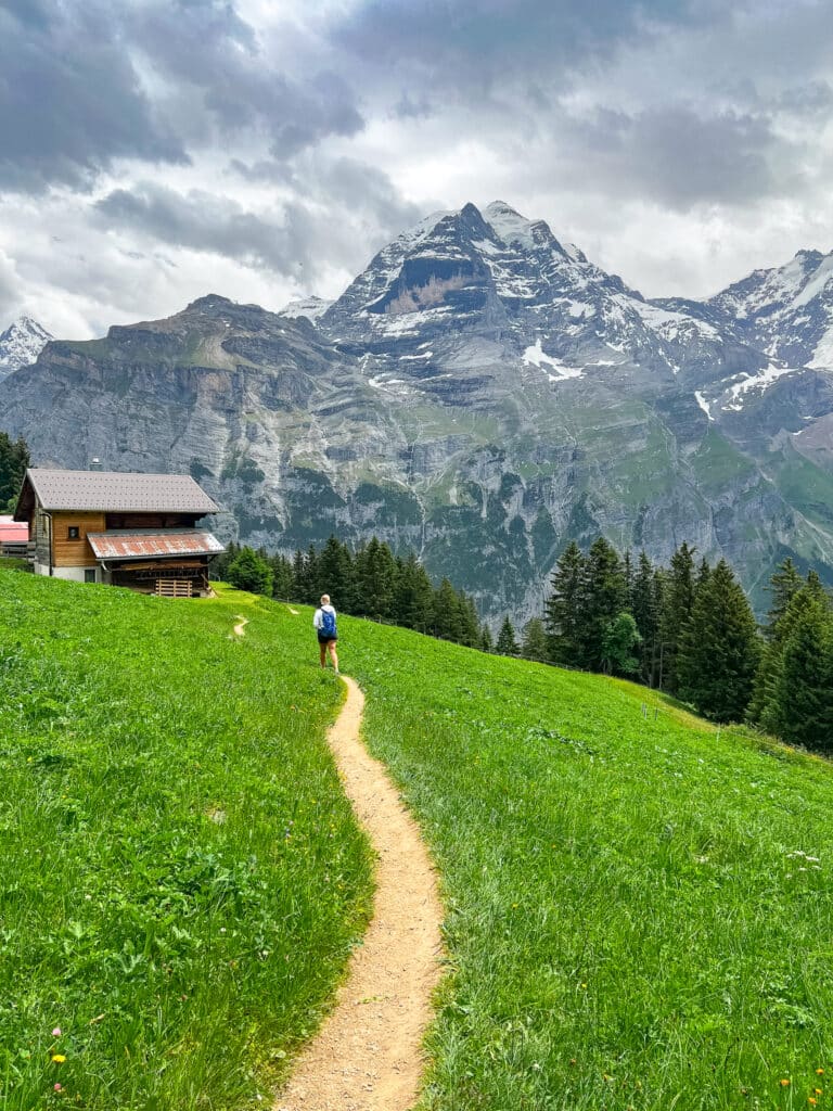 Hiker following a narrow dirt trail toward a wooden alpine chalet with a snow-capped peak behind, on the Mürren Northface Trail