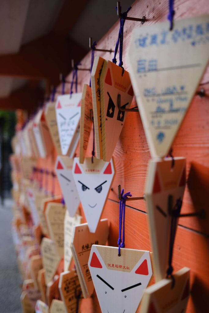 Close-up of wooden fox-shaped ema plaques with handwritten wishes and drawings, hanging in rows at a shrine in Japan. The fox faces have unique expressions, and the background shows an orange wooden structure.