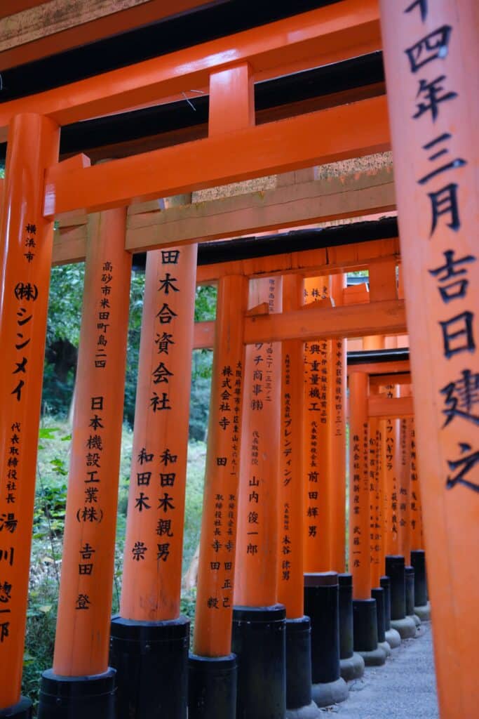 A perspective view through a tunnel of vibrant orange torii gates at Fushimi Inari Shrine in Kyoto, Japan, with black Japanese calligraphy inscriptions on each gate and a gravel path beneath.