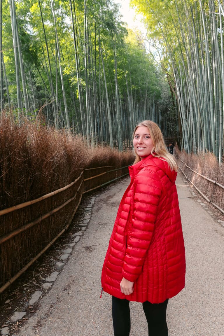 Woman in a red coat standing on the path through Kyoto’s Arashiyama Bamboo Forest, surrounded by tall bamboo stalks in soft morning light