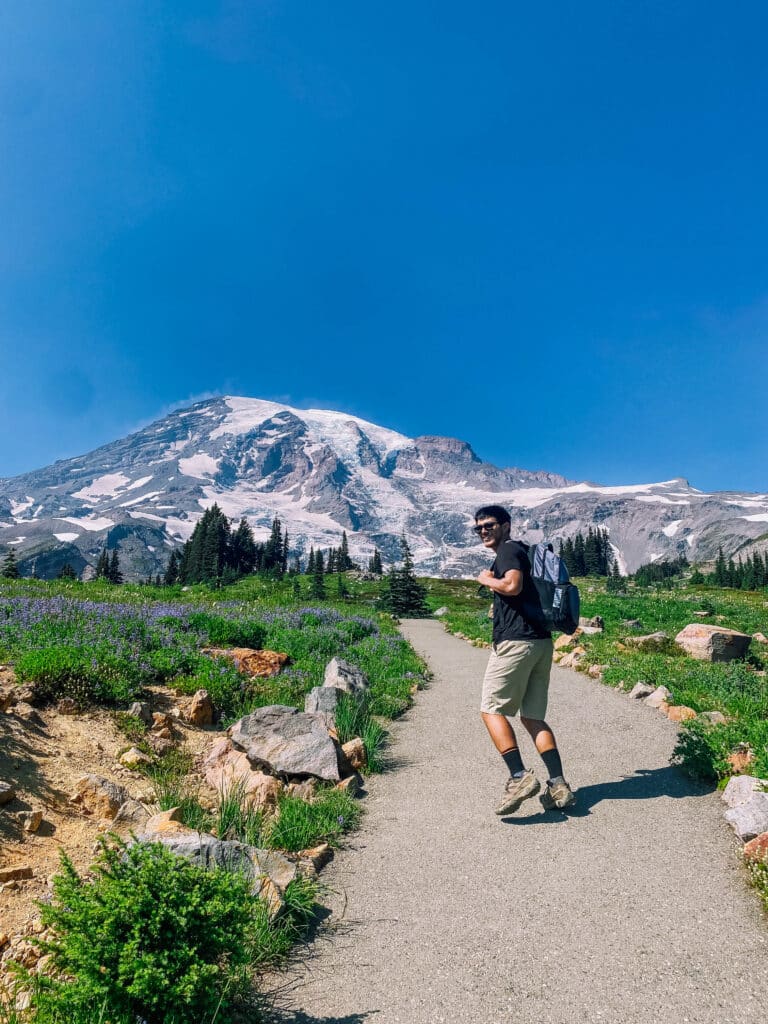 Back view of a hiker approaching Mount Rainier on a wildflower-lined trail in Paradise