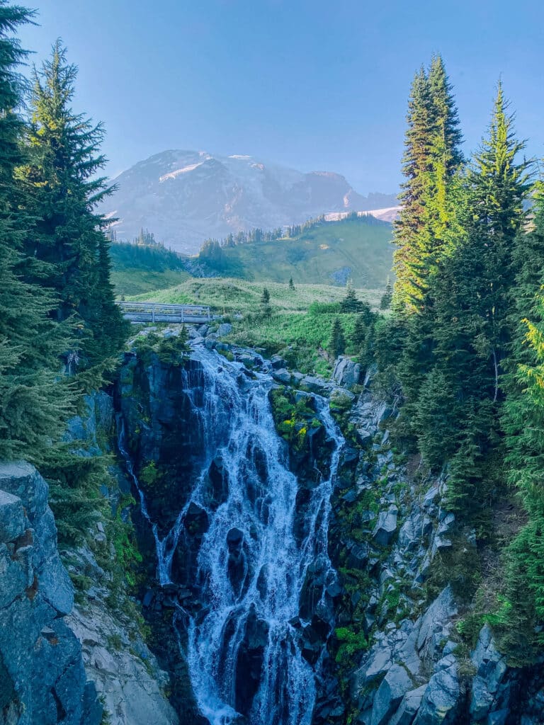 Myrtle Falls cascading through a rocky gorge with dense evergreens and a peek of Mount Rainier in the background