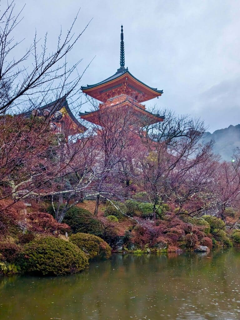 A Three-story pagoda surrounded by cherry blossoms and a peaceful pond, partially obscured by trees at Kiyomizu-dera in Eastern Kyoto