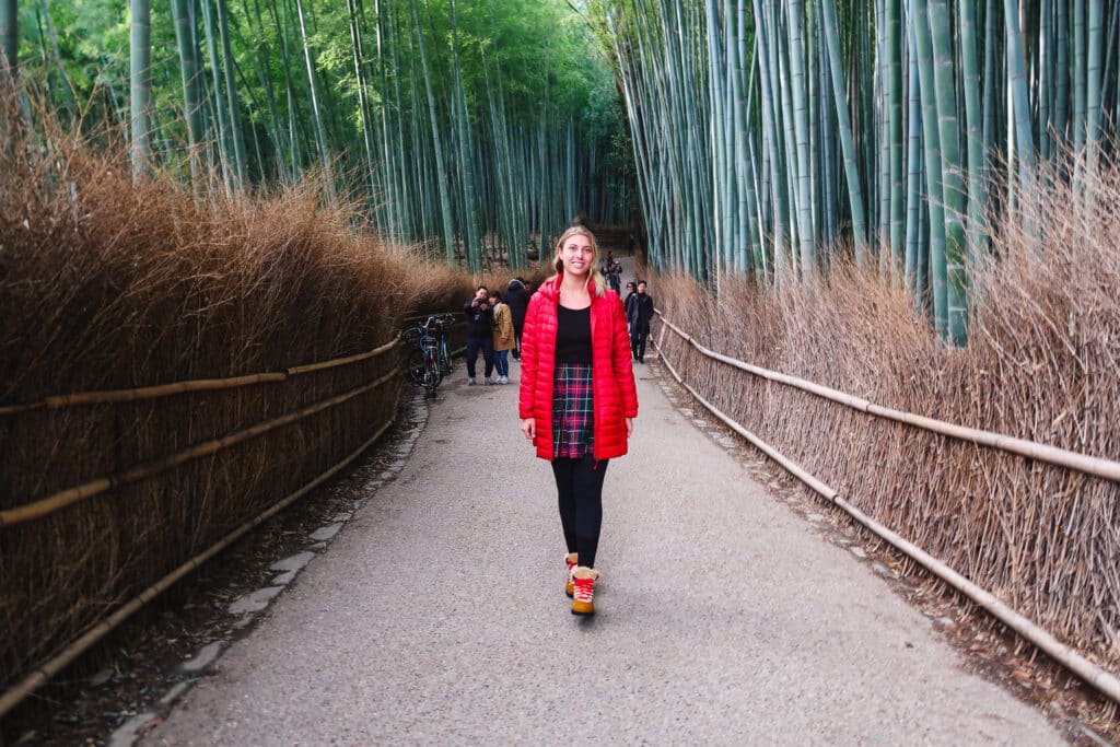 "Woman walking down the central path of Arashiyama Bamboo Forest in Kyoto, surrounded by tall bamboo and winter brush on a quiet morning."