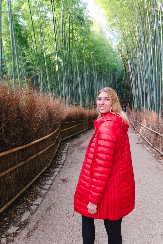 Woman in a red coat standing on the path through Kyoto’s Arashiyama Bamboo Forest, surrounded by tall bamboo stalks in soft morning light