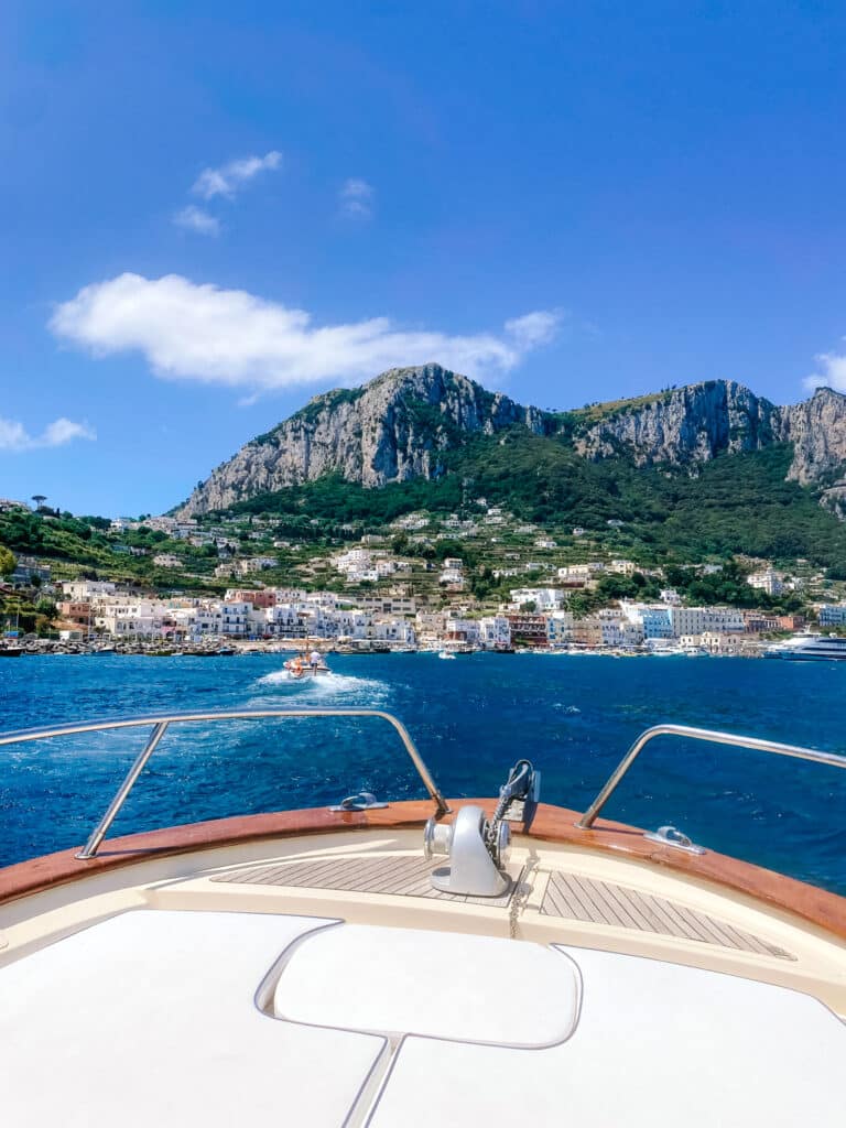 The colourful waterfront buildings of Marina Grande coming into view over the boat's bow, with Monte Solaro and Capri's dramatic mountain ridge towering above.