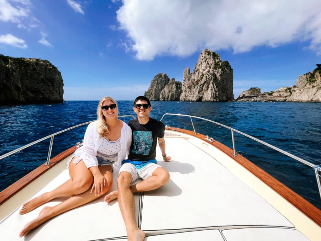A couple posing together on the bow of the boat with all three Faraglioni rocks perfectly framed behind them against a bright blue sky.