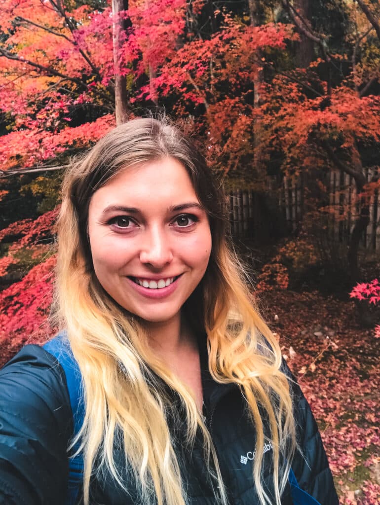 A visitor in a black puffer coat taking a selfie with a red japanese maple tree