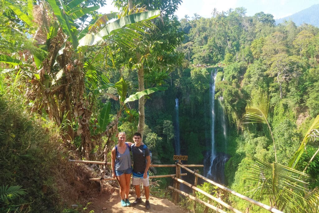 Two travelers by a yellow wooden “Sekumpul” sign pointing toward the waterfall, with the twin falls visible in the distance.