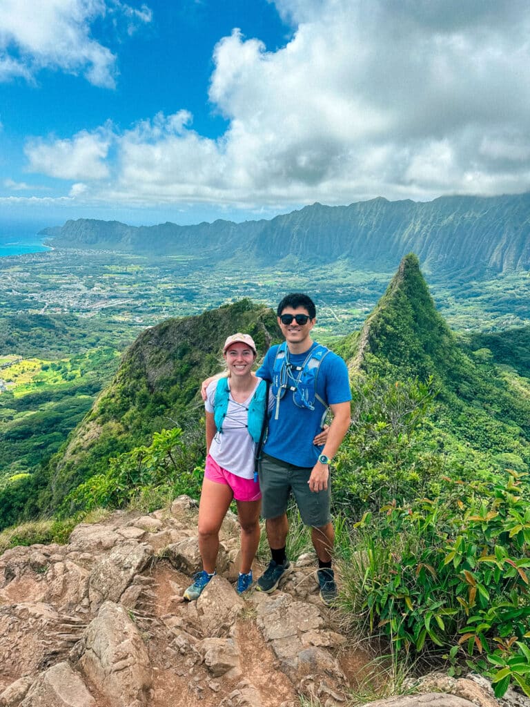 Two hikers standing at a narrow viewpoint on Olomana with steep ridges and valley views behind them.