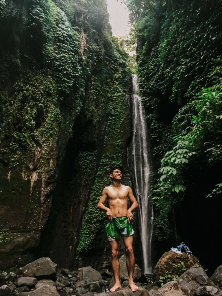 Traveler standing shirtless in front of a narrow jungle waterfall, surrounded by towering green walls in Sekumpul valley.