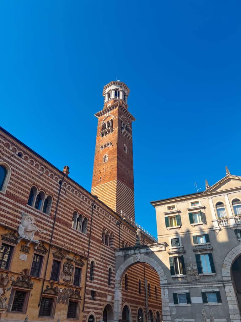 Torre dei Lamberti seen from below, framed by historic buildings and a clear blue sky.