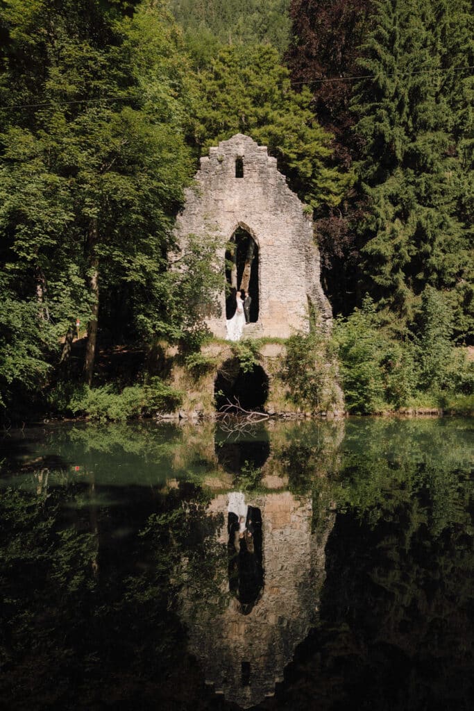Bride and groom reflected in a tranquil mountain lake with a stone chapel ruin in the background. Photo credit: Mathilde Rietsch