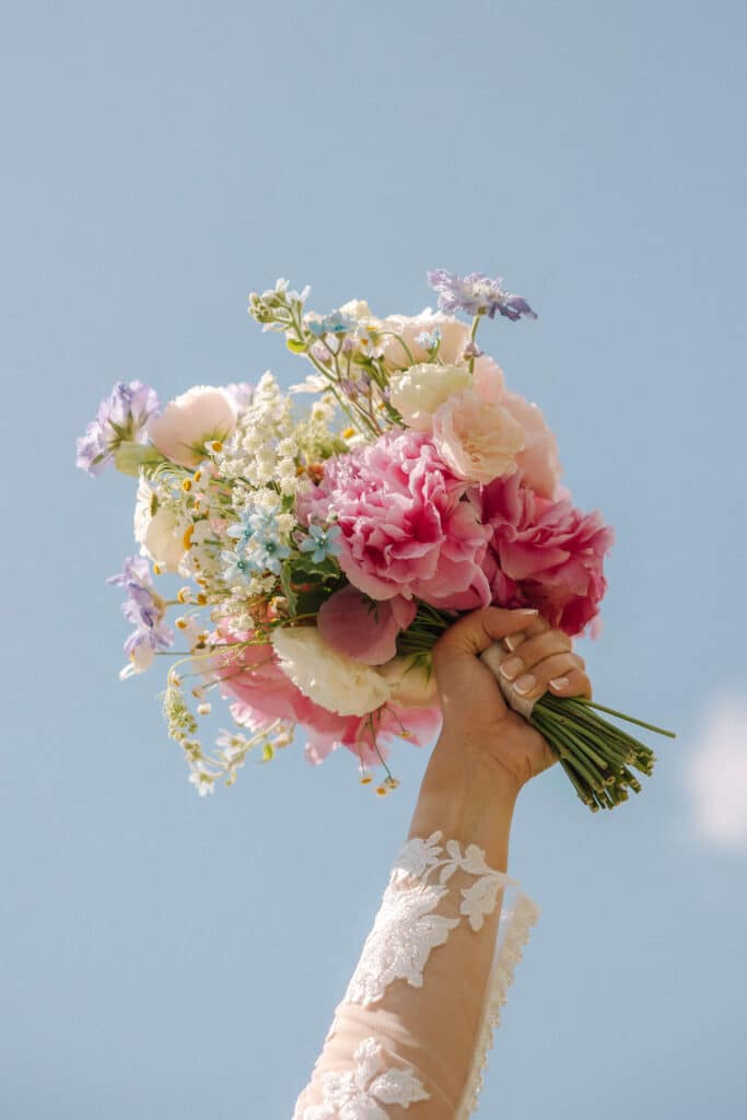Bride raising her wildflower bouquet triumphantly against a clear blue sky, showing off pink peonies and blue flowers that are reminiscent of forget-me-nots. Photo credit: Mathilde Rietsch