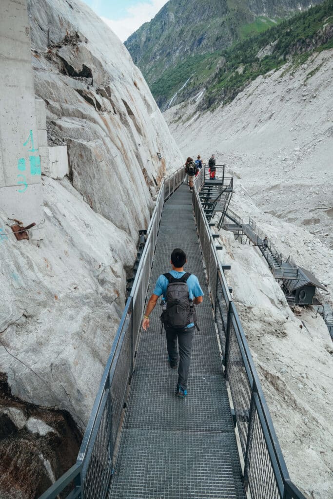 Tourists descending steep metal stairs toward the glacier, with rocky terrain in view.