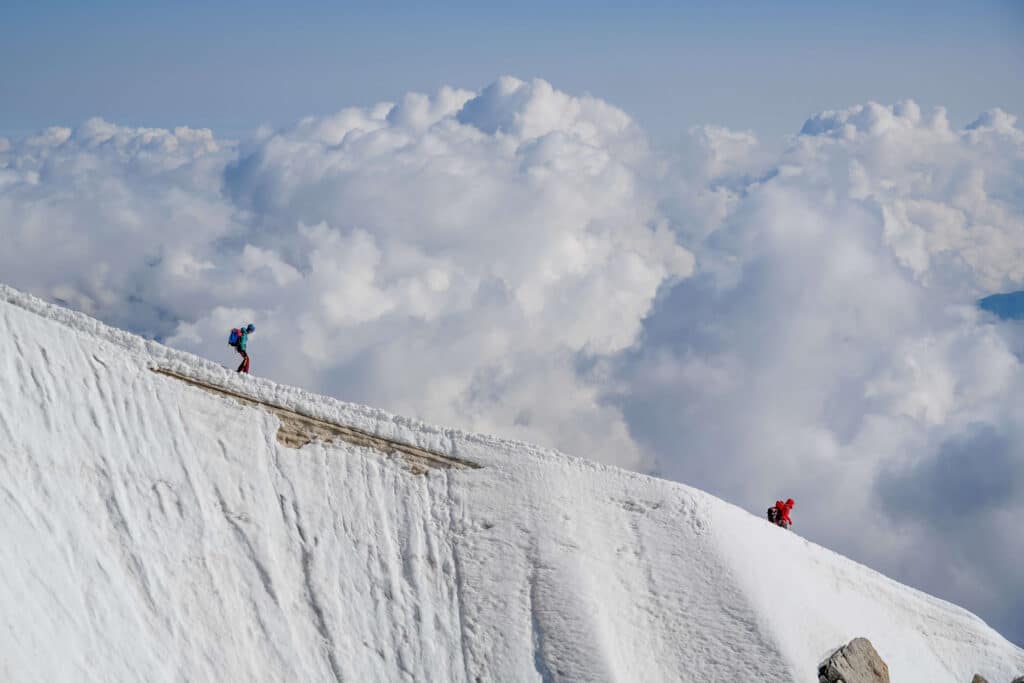 Two alpinists standing on narrow snow ridge high above the valley with puffy clouds in the background