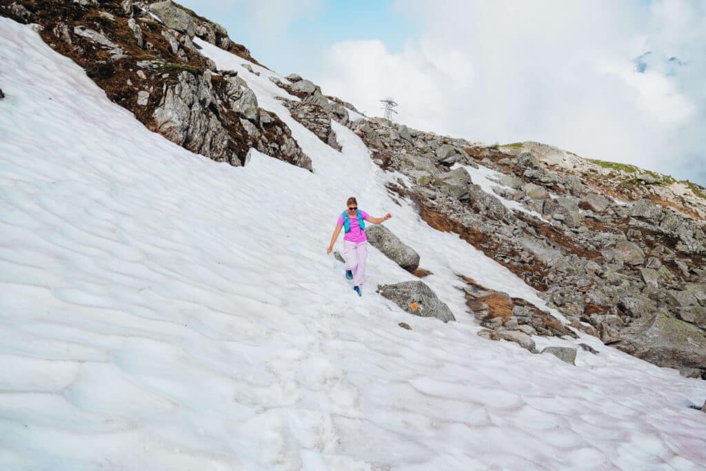 Hiker crossing a snowfield near Lac Bleu, surrounded by rocky alpine terrain and soft clouds.