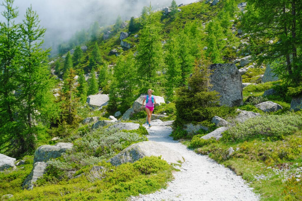 Trail winds through green larches and mist on the Grand Balcon Nord, with a trail runner ahead on the path.