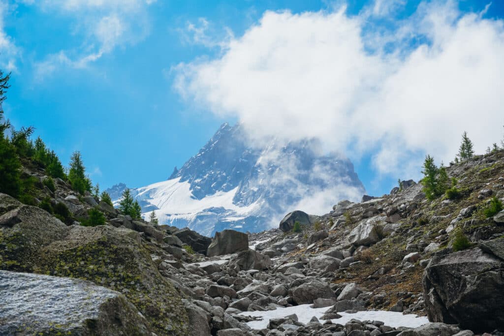 View of the Aiguille du Midi and glacier above a rocky trail framed by larches and wild terrain.
