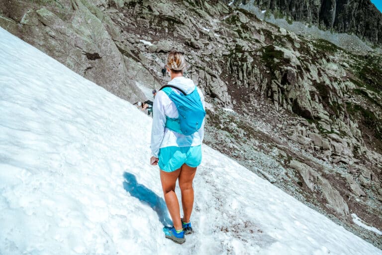 Back view of a hiker in a pastel windbreaker and trail runners walking across a snowy traverse in Chamonix, illustrating early summer snow conditions on high-elevation trails.