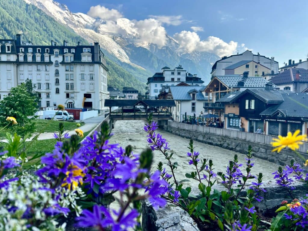 Colorful wildflowers in the foreground of Chamonix’s town center, with white alpine buildings lining a glacial river and Mont Blanc towering in the background.