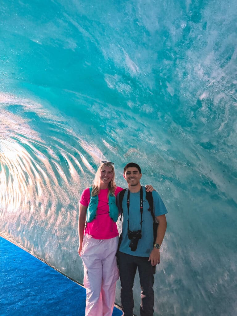 Two visitors inside the Mer de Glace Grotto glacier cave, surrounded by shimmering blue ice walls.