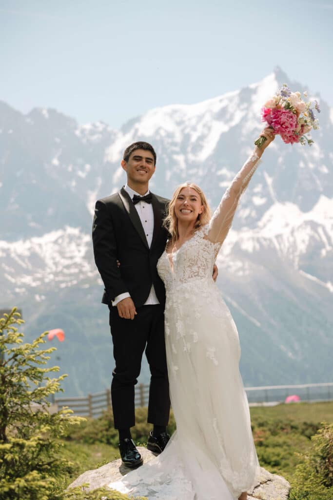 Bride and groom standing on a rock at Planpraz, with the bride raising her bouquet triumphantly and Mont Blanc glowing in the background. Photo credit: Mathilde Rietsch.