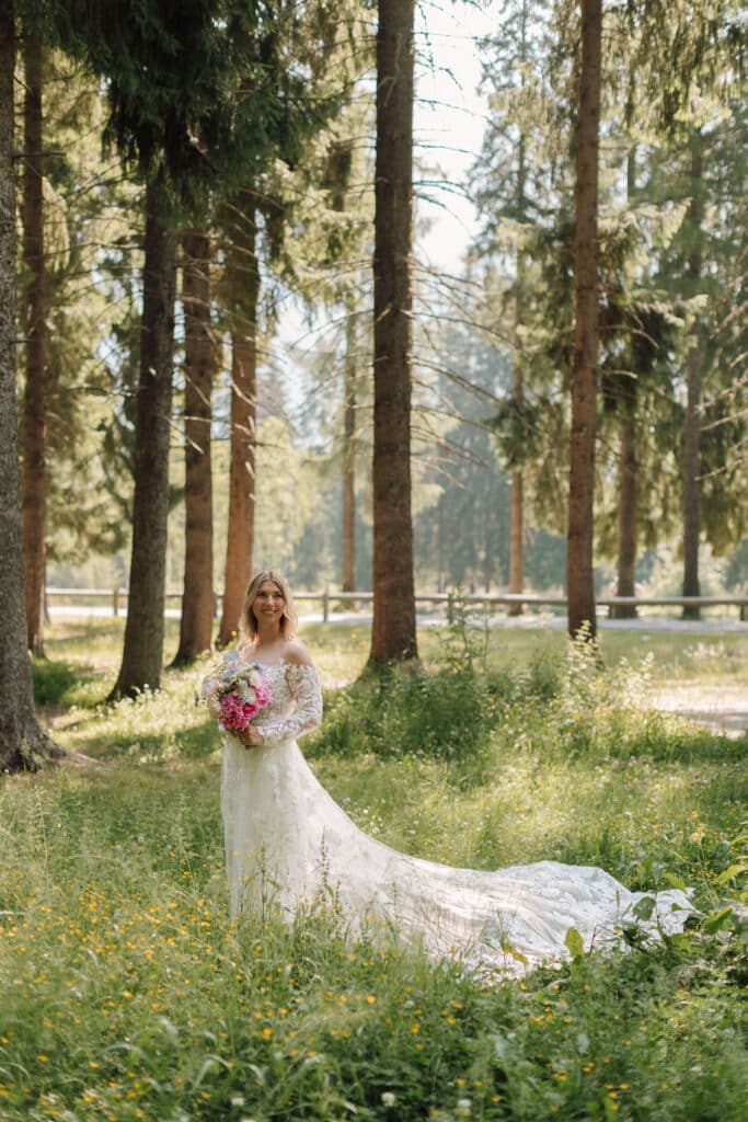 Bride standing in a sun-dappled wildflower meadow at Les Gaillands, holding a pink and blue bouquet with her lace train trailing behind her in the grass. Photo credit: Mathilde Rietsch