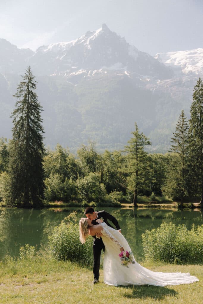 Newlyweds sharing a joyful kiss in front of a forested lake at Les Gaillands. Photo credit: Mathilde Rietsch.