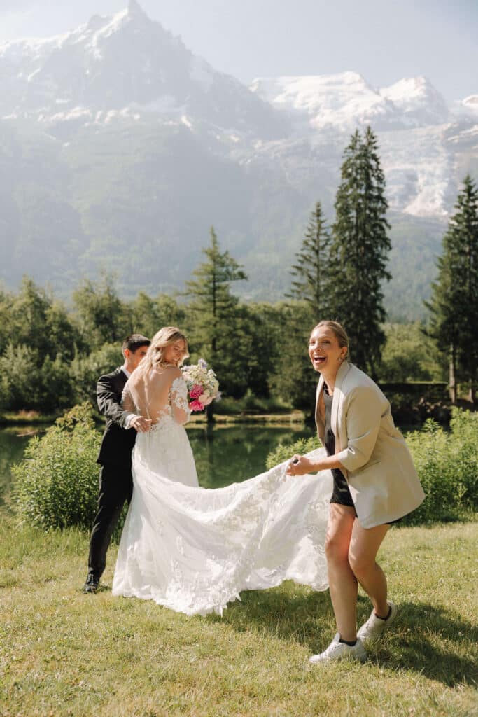 Bride smiling while Laura Wilson-North helps adjust her train, with the groom standing nearby and Aiguille du Midi in the background. Photo credit: Mathilde Rietsch
