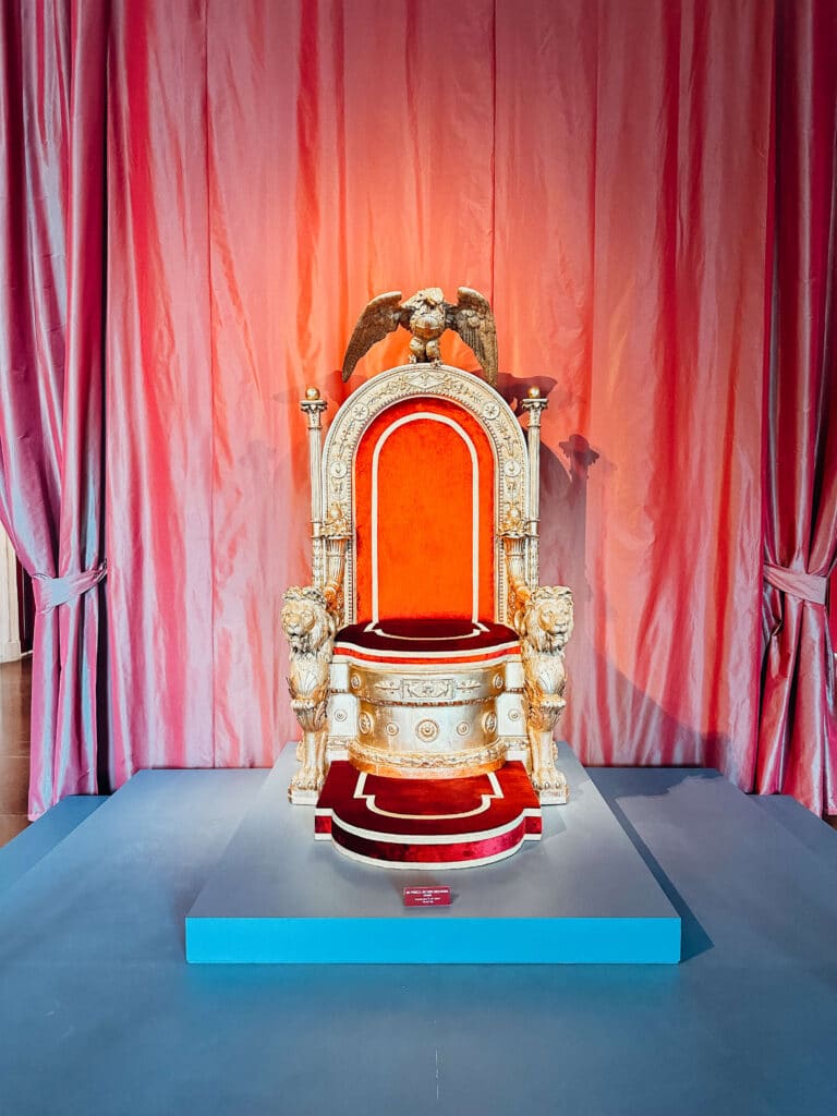 A dramatic red velvet throne set against pink curtains under spotlight in the Venaria Reale