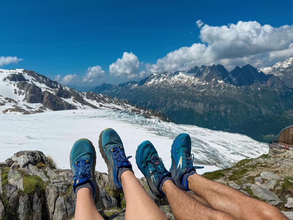 wo pairs of hiking shoes dangling over a rocky ledge, overlooking the snow-covered Albert 1er Glacier and the Mont Blanc range in the distance.