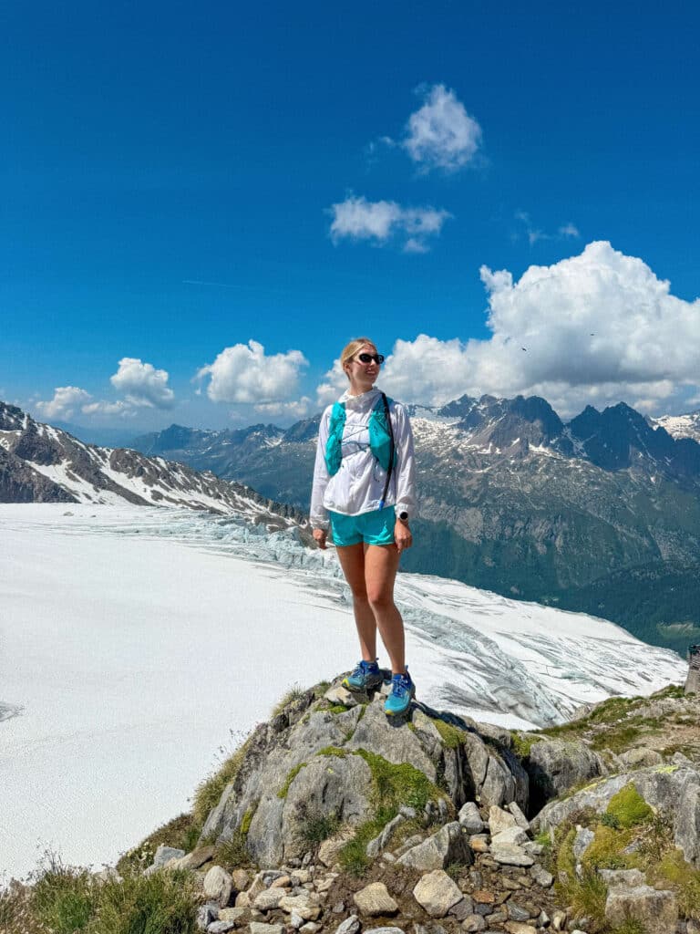 Hiker standing on a rocky outcrop above the Albert 1er Glacier, wearing a turquoise cap and blue trail shoes, with sweeping views of the Mont Blanc massif behind.