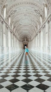Person in a blue shirt walking through a long marble gallery with black and white checkered floor and arched ceilings at Venaria Reale