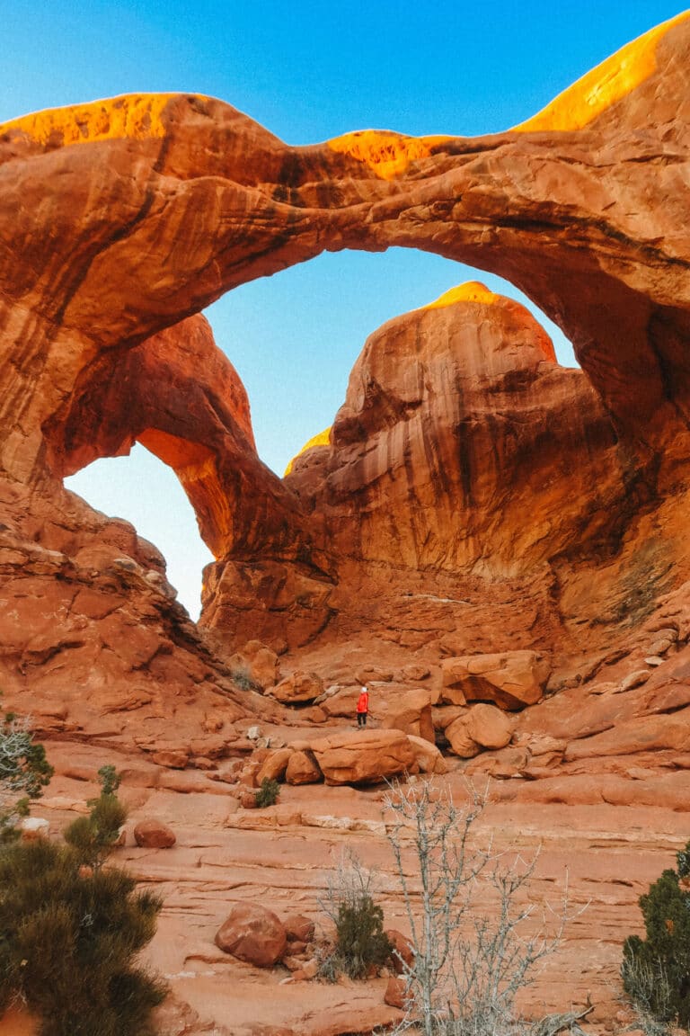Head-on view of Double Arch with deep blue sky above, with a little figure in red standing on a rock below the arms of the arches